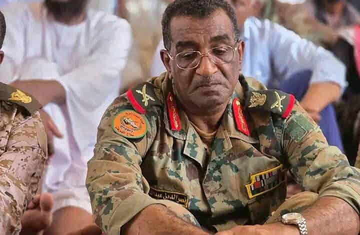 قائد عسكري سوداني في اجتماع عسكري وسط حضور جماهيري، يرتدي زي الجيش السوداني، ويظهر على وجهه التركيز والانتباه، في حدث مهم يخص الشأن السوداني.